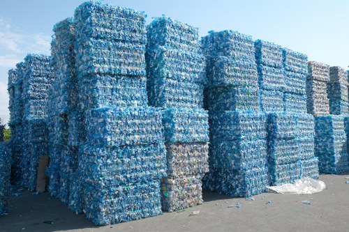A large outdoor storage area features multiple stacks of compacted plastic bottles, predominantly blue with some clear and green bottles visible, arranged in tall, rectangular bales on the paved ground. The bales are tightly packed, with some evidence of small loose bottles or debris scattered around the base. In the background, the sky is clear and blue, indicating bright daytime conditions. The scene is typical of a waste management facility or recycling centre, where plastic bottles are prepared for disposal or recycling. The presence of these compressed bales of plastic bottles aligns with rubbish removal services such as those offered by Commercial Waste Removal Sudbury, which handle bulk waste materials efficiently. The environment appears clean and organized, emphasizing the importance of proper rubbish collection and recycling practices in areas like Sudbury, UK, ensuring local waste management standards are met effectively.