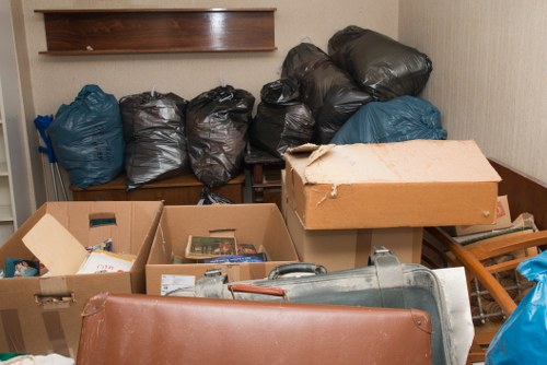 A collection of black and blue garbage bags filled with waste are stacked along a wooden shelf or bench in an indoor storage area, with some bags leaning against the wall and others placed atop or beside cardboard boxes filled with miscellaneous papers and packaging materials. A large, partially opened cardboard box reveals various waste items inside, while an older, brown leather chair or seat is positioned in the foreground, partially obstructing the view of a metal object, possibly a vacuum cleaner or industrial equipment, lying on the floor. To the right, a small wooden folding chair is visible, and the background shows plain beige walls with a wooden shelf mounted above, suggesting a makeshift storage space or warehouse environment. The scene appears cluttered but organized for waste collection, relevant to services like those offered by Commercial Waste Removal Sudbury for rubbish clearance in the local area.