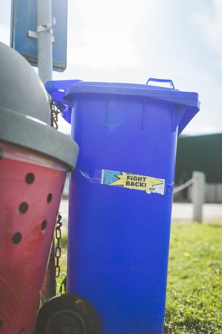 A middle-aged man with dark hair, dressed in a black T-shirt with white text and dark trousers, is seen leaning over a modern, metallic, cylindrical rubbish bin with two compartments, positioned on a