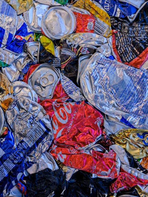 A close-up view of a pile of crushed and crumpled aluminium beverage cans, predominantly silver with some red, blue, black, and yellow packaging labels, scattered on a surface possibly outdoors or in 